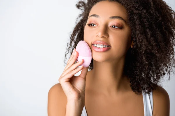 Sorrindo menina afro-americana com aparelho dentário usando escova facial de limpeza de silicone, isolado em cinza — Fotografia de Stock