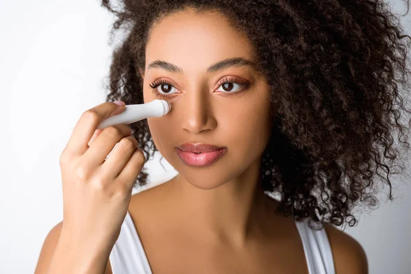 Привлекательная африканская американка с помощью увлажняющего глазного ролика, изолированного на сером — стоковое фото