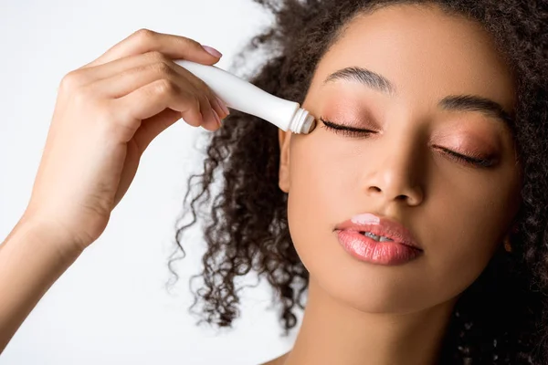 Atractiva chica afroamericana con los ojos cerrados usando rodillo de ojo hidratante, aislado en gris - foto de stock