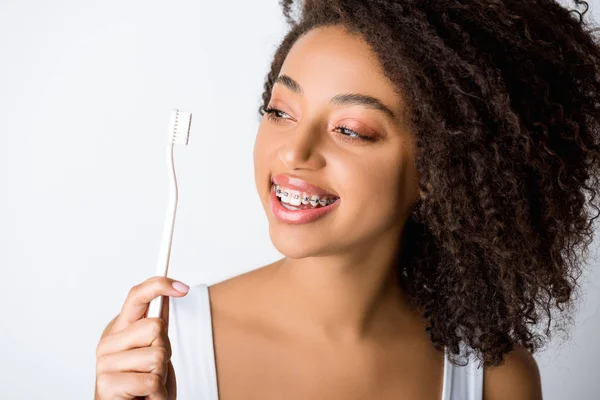 Smiling african american woman with dental braces looking at toothbrush, isolated on grey — Stock Photo