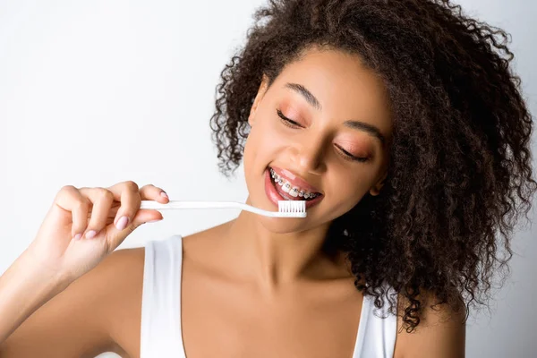Cheerful african american woman with dental braces looking at toothbrush, isolated on grey — Stock Photo