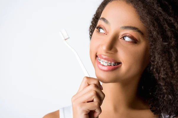 Belle femme afro-américaine bouclée avec des appareils dentaires regardant la brosse à dents, isolée sur gris — Photo de stock