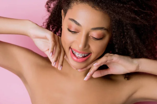 Atractiva chica afroamericana desnuda sonriente con frenos dentales, aislado en rosa - foto de stock