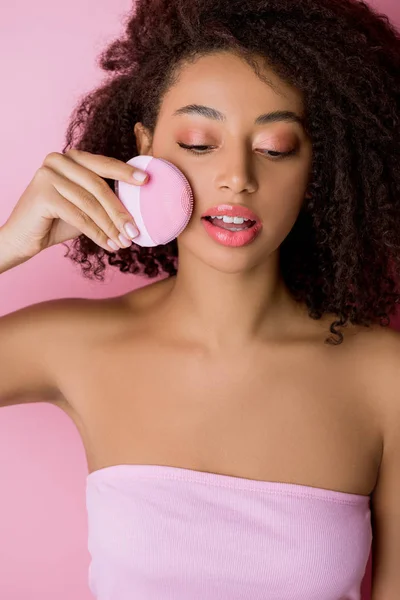 Afro-américaine les yeux fermés à l'aide d'une brosse nettoyante au silicone, isolée sur rose — Photo de stock