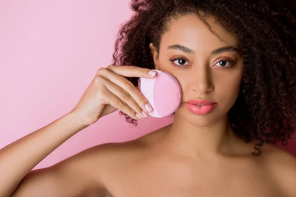 Chica afroamericana atractiva desnuda con los ojos cerrados usando cepillo facial de limpieza de silicona, aislado en rosa - foto de stock