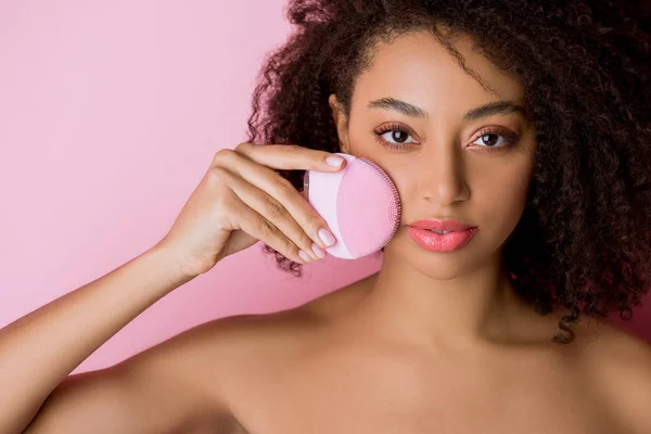 Chica afroamericana atractiva desnuda con los ojos cerrados usando cepillo facial de limpieza de silicona, aislado en rosa — Stock Photo