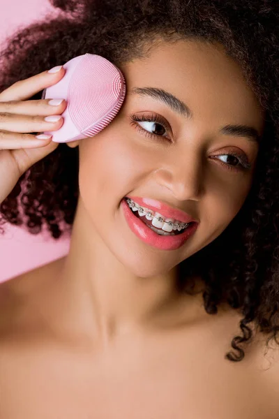 Mujer afroamericana con los ojos cerrados usando cepillo facial limpiador de silicona, aislado en rosa — Stock Photo