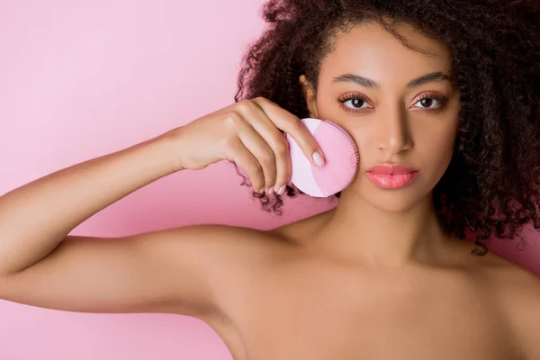 Mujer afroamericana desnuda con los ojos cerrados usando cepillo facial limpiador de silicona, aislado en rosa - foto de stock