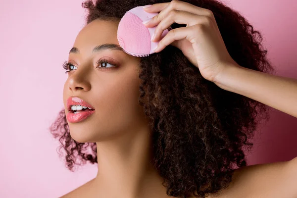 Mujer afroamericana con los ojos cerrados usando cepillo facial limpiador de silicona, aislado en rosa — Stock Photo