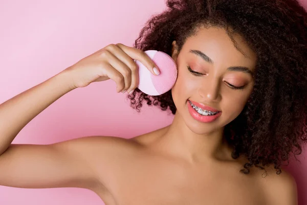 Mulher americana africana nua com olhos fechados usando escova facial de limpeza de silicone, isolada em rosa — Fotografia de Stock