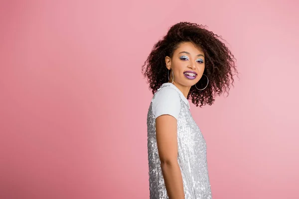 Cheerful stylish african american girl with dental braces, with silver glitter eyeshadows and purple lips dancing in paillettes dress, isolated on pink — Stock Photo