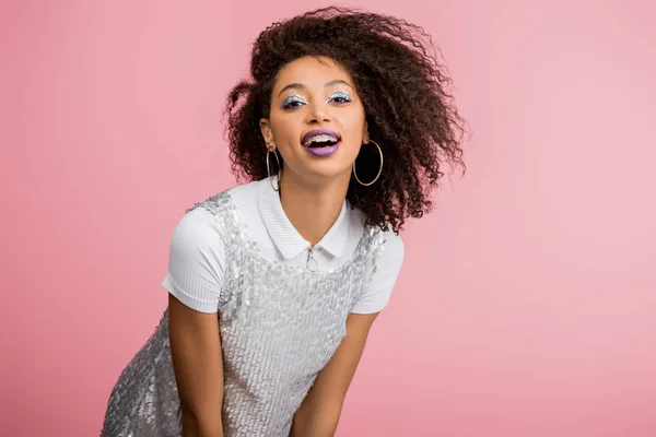 Elegante ragazza afroamericana sorridente con bretelle dentali, con ombretti glitter argento e labbra viola che ballano in abito paillettes, isolato su rosa — Foto stock