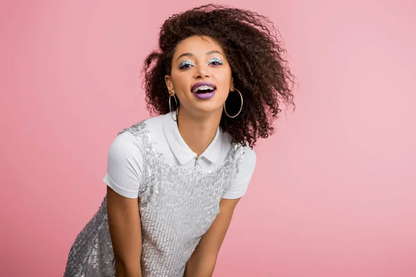 Elegante chica afroamericana sonriente con frenos dentales, con sombras de ojos de brillo plateado y labios morados bailando en paillettes vestido, aislado en rosa - foto de stock