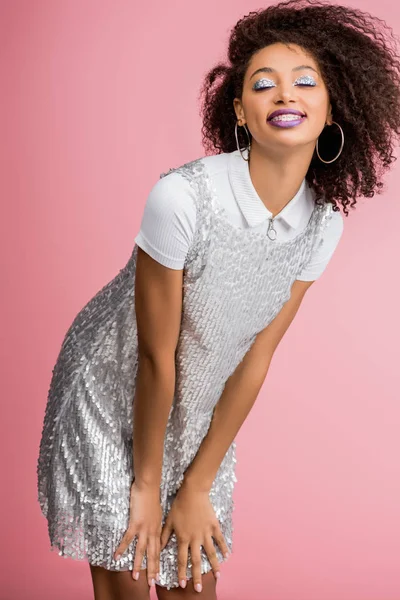 Улыбающаяся африканская американка с зубными брекетами, с серебряными блестящими тенями для век и фиолетовыми губами, танцующими в платье из пайетт, изолированные на розовом — стоковое фото