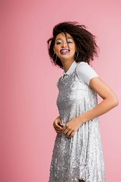 Jolie fille afro-américaine gaie avec des appareils dentaires, avec des fards à paupières à paillettes argentées et des lèvres violettes dansant en robe paillettes, isolée sur rose — Photo de stock