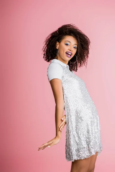 Happy african american girl with dental braces, with silver glitter eyeshadows and purple lips dancing in paillettes dress, isolated on pink — Stock Photo