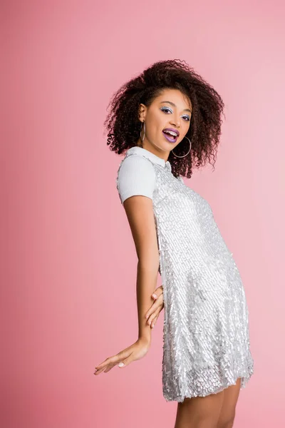 Heureuse fille afro-américaine avec des bretelles dentaires, avec des fards à paupières à paillettes argentées et des lèvres violettes dansant en robe paillettes, isolée sur rose — Photo de stock