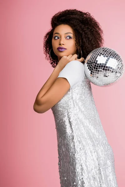 Elegante ragazza afroamericana in paillettes vestito tenendo palla disco, isolato su rosa — Foto stock