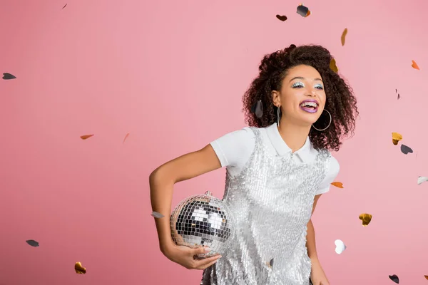 Sorrindo menina americana africana em paillettes vestido segurando bola de discoteca, isolado em rosa com confete — Fotografia de Stock