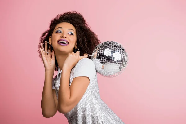 Smiling african american girl in paillettes dress holding disco ball, isolated on pink with confetti — Stock Photo