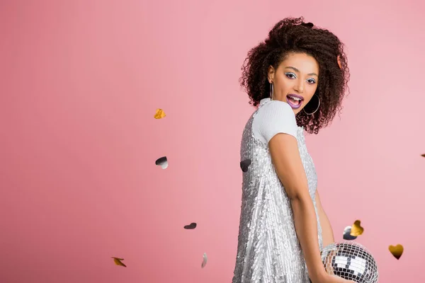 Cheerful african american girl in paillettes dress holding disco ball, isolated on pink with confetti — Stock Photo