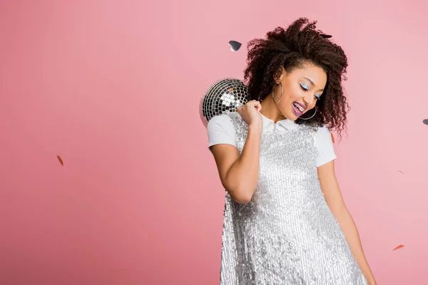 Smiling african american girl in paillettes dress holding disco ball, isolated on pink with confetti — Stock Photo