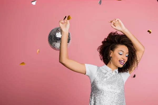 Счастливая привлекательная африканская американская девушка в пайетках платье танцы с дискотекой мяч, изолированные на розовый с конфетти — стоковое фото