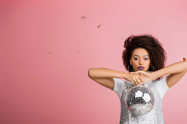 Attraente ragazza afroamericana in paillettes vestito tenendo palla disco, isolato su rosa con coriandoli — Foto stock