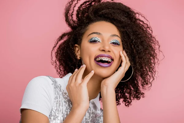 Excitée afro-américaine avec des bretelles dentaires, avec des fards à paupières à paillettes argentées et des lèvres violettes portant une robe paillettes, isolée sur rose — Photo de stock