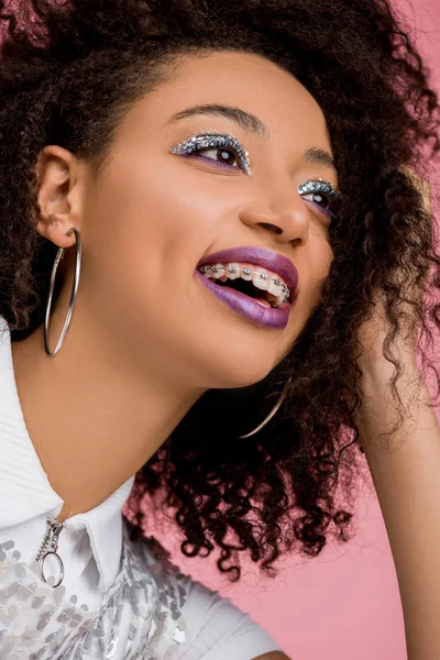 Joyeuse fille afro-américaine avec des bretelles dentaires, avec des fards à paupières à paillettes argentées et des lèvres violettes portant une robe paillettes, isolée sur rose — Photo de stock