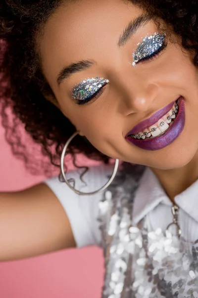 Felice ragazza afroamericana con bretelle dentali, con ombretti scintillanti d'argento e labbra viola che indossano un abito paillettes, isolato su rosa — Foto stock