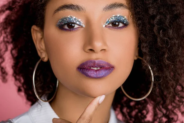 Atractiva chica afroamericana con sombras de ojos de brillo plateado y labios morados, aislado en rosa - foto de stock