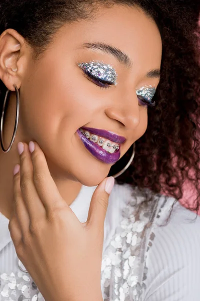 Joyeuse fille afro-américaine avec des fards à paupières à paillettes argentées et des lèvres violettes portant une robe paillettes, isolée sur rose — Photo de stock