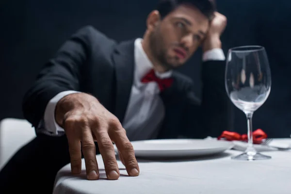Enfoque selectivo del hombre elegante con la mano por la cabeza sentado en la mesa servida aislado en negro - foto de stock