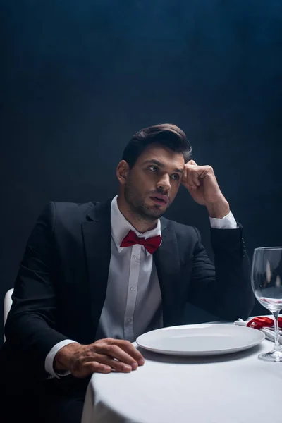 Bello uomo in giacca e cravatta guardando lontano al tavolo servito isolato su nero — Foto stock