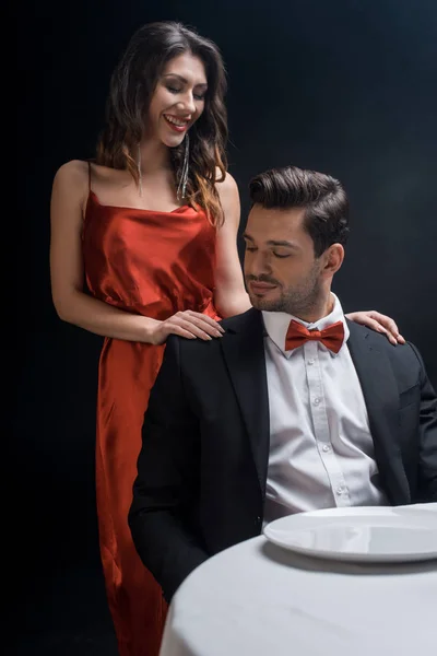 Mujer sonriente en vestido elegante de pie por un hombre guapo en la mesa servida aislado en negro - foto de stock