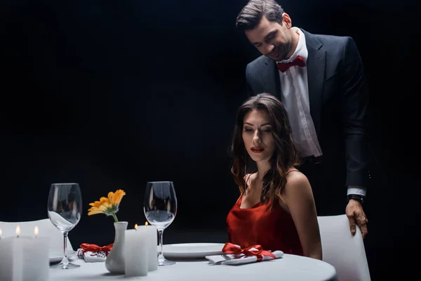 Hombre guapo de pie detrás de la mujer elegante en la mesa servida aislado en negro - foto de stock