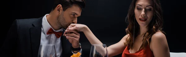 Panoramic shot of handsome man kissing hand of elegant woman by wine glasses isolated on black — Stock Photo