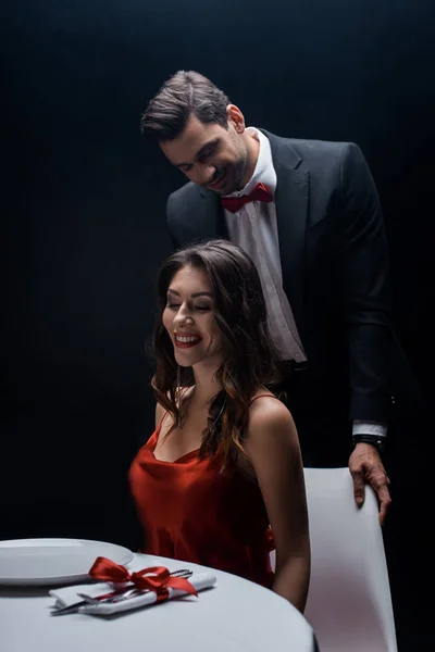 Elegante hombre de pie detrás de la hermosa mujer sonriente en la mesa servida aislado en negro - foto de stock