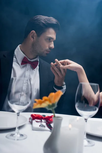 Vista lateral del hombre guapo sosteniendo la mano de la mujer y mirando a la mesa servida sobre fondo negro con humo - foto de stock