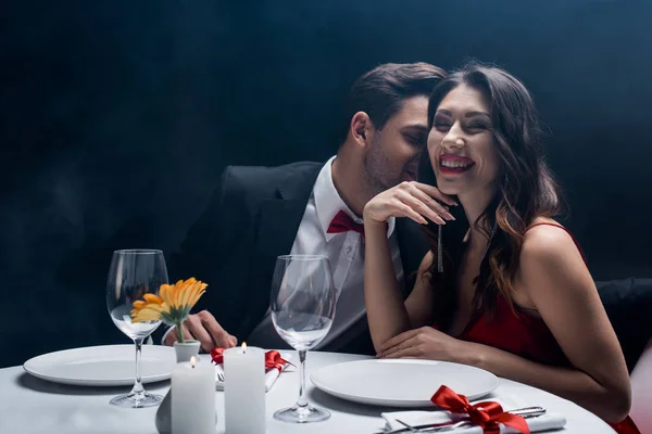 Elegant man whispering to smiling woman during romantic dinner on black background with smoke — Stock Photo