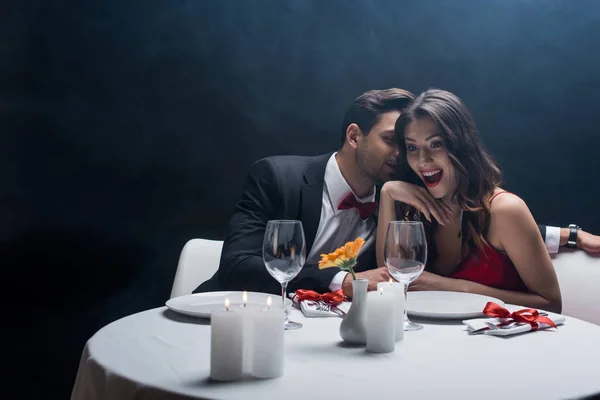 Handsome man in suit whispering to shocked woman at served table on black background with smoke — Stock Photo