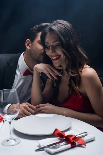 Hombre guapo susurrando a hermosa mujer sonriente en la mesa servida sobre fondo negro con humo - foto de stock