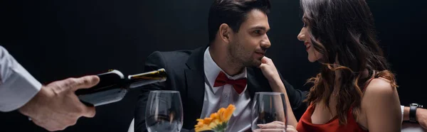 Enfoque selectivo de la pareja sonriente en la mesa junto al camarero vertiendo vino en vasos aislados en negro - foto de stock