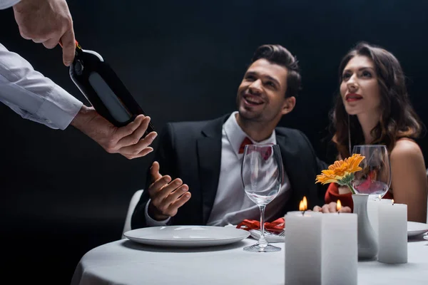 Enfoque selectivo de camarero mostrando botella de vino a elegante pareja en mesa servida aislada en negro - foto de stock