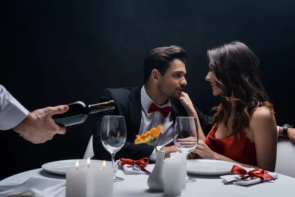 Enfoque selectivo de pareja durante la cena romántica y camarero vertiendo vino tinto aislado en negro - foto de stock