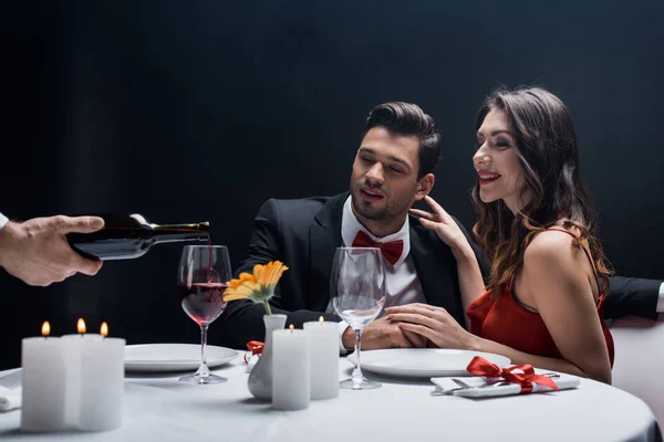 Garçom derramando vinho em vidro com casal elegância na mesa servida isolado em preto — Fotografia de Stock