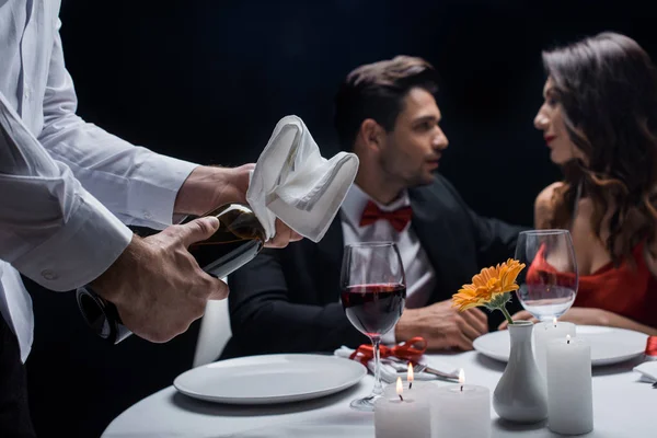 Enfoque selectivo del camarero con botella de vino y toalla y pareja de elegancia en la mesa servida en el fondo aislado en negro - foto de stock