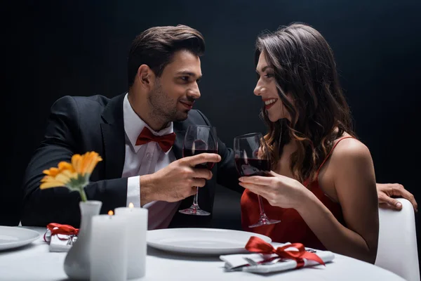 Vista lateral de pareja sonriente con copas de vino mirándose a la mesa servida aislada en negro - foto de stock