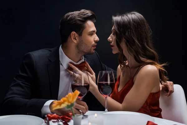 Vista lateral de la hermosa mujer ajustando la pajarita del hombre guapo durante la cena romántica aislada en negro - foto de stock